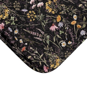 K&ouml;nnte beinhalten: Eine schwarze Badematte mit einem Blumenmuster, das verschiedene Blumen in den Farben Rosa, Gelb und Lila zeigt. Die Blumen sind in einem zuf&auml;lligen Muster auf einem dunklen Hintergrund angeordnet.