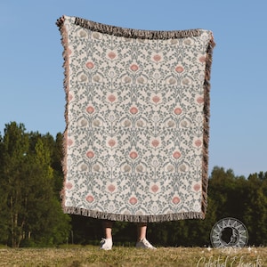 May include: A woven throw blanket with a floral pattern in shades of pink, green, and white. The blanket has a fringed edge and is being held up by a person in front of a blue sky and green trees.