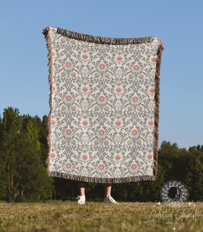 May include: A woven throw blanket with a floral pattern in shades of pink, green, and white. The blanket has a fringed edge and is being held up by a person in front of a blue sky and green trees.