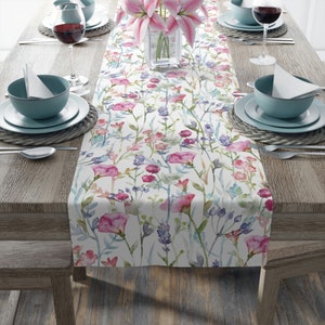 May include: A white table runner with a watercolor floral pattern featuring pink, purple, and green flowers. The runner is laid out on a wooden table with two place settings on each side.