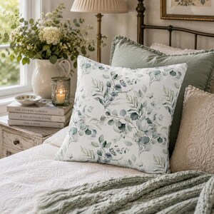 May include: A square throw pillow featuring a white background with a repeating pattern of green and blue-toned eucalyptus leaves and branches. The pillow is positioned on a bed alongside a solid sage green pillow and a textured beige pillow.