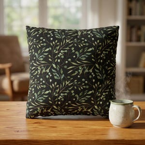 May include: Square throw pillow with a black background and a green and white botanical pattern. A small, off-white ceramic mug with a green rim sits on a wooden table in front of the pillow.