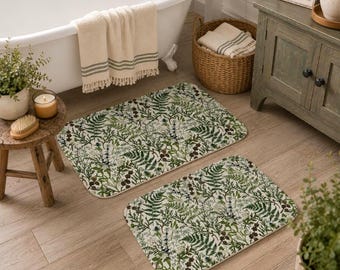 May include: Two rectangular bathroom rugs featuring a detailed botanical print of green ferns, dark brown pinecones, and small blue flowers on a white background. The rugs have rounded corners and a soft, flat-weave texture for bathroom floors.