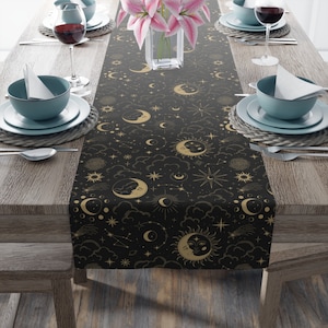 May include: A black table runner with a gold celestial pattern featuring moons, stars, and clouds. The runner is on a wooden table with place settings.
