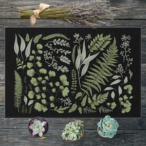 May include: A rectangular black doormat featuring a botanical design with various green and white leaves and ferns. A bundle of dried lavender with a burlap bow sits above the mat. Three potted succulents are at the bottom.