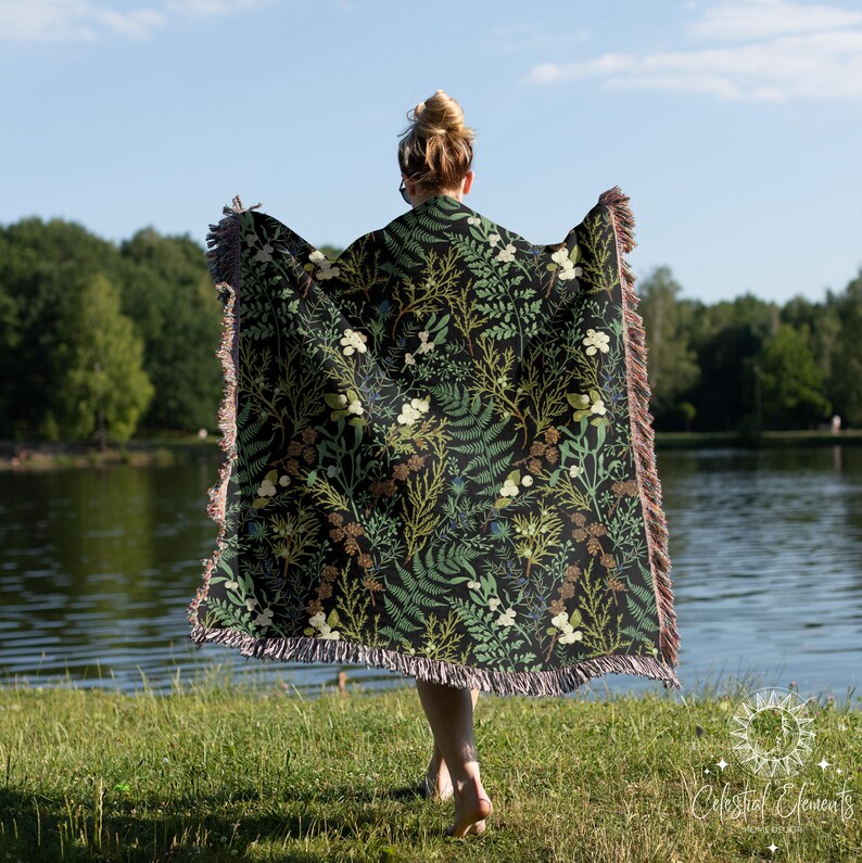 May include: A woven throw blanket with a dark green, black, and white floral pattern. The blanket has a fringed edge and is being held by a person standing in a grassy field.