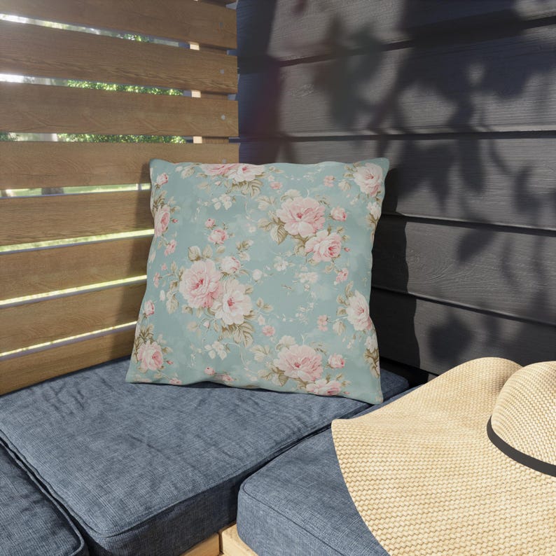 May include: Square throw pillow with a floral pattern of pink roses on a light blue background. The pillow is placed on a blue cushion, with a straw hat in the foreground. The pillow adds a touch of elegance to outdoor seating.