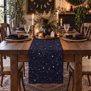 May include: A dark navy blue table runner featuring a white celestial pattern of scattered stars and constellations. The runner is laid flat across a wooden dining table, centered beneath a decorative centerpiece with candles and a black vase.