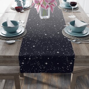 May include: A black table runner with a white star pattern. The runner is on a wooden table with two place settings on each side. The place settings include a blue bowl, a white napkin, a glass of red wine, and silverware.