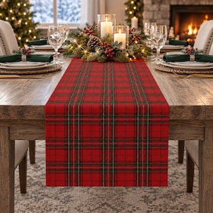 May include: A rectangular table runner featuring a red, green, and white plaid pattern. The fabric is laid flat across the center of a wooden dining table, extending from the foreground toward a centerpiece of candles and greenery.