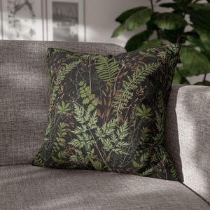 May include: Square throw pillow with a dark background and a botanical pattern of green and brown leaves. The pillow is resting on a gray sofa, creating a contrast between the dark pillow and the light-colored furniture. The pillow adds a touch of nature to the room.