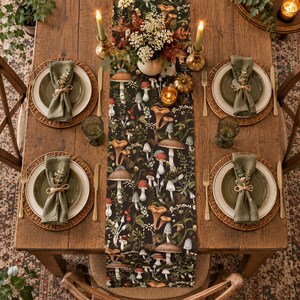 May include: A wooden dining table set for four with a black table runner featuring a colorful mushroom and botanical print. Each place setting includes a woven placemat, a white plate, a green plate, a green napkin, and a gold-toned flatware set.
