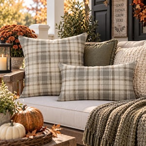 May include: A collection of decorative throw pillows featuring a beige and gray plaid pattern, including one square pillow and one rectangular lumbar pillow, arranged on a light-colored outdoor bench with a textured olive green pillow.