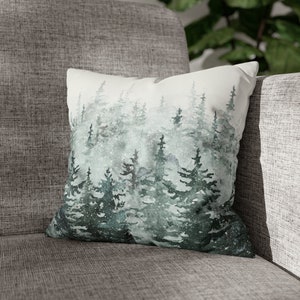 May include: Square decorative pillow featuring a watercolor forest scene in shades of green and gray. The design depicts evergreen trees with a snowy effect, creating a serene, natural aesthetic. The pillow is placed on a gray upholstered chair.
