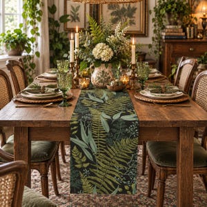 May include: A long rectangular table runner featuring a dark background with a detailed botanical print of various green ferns and foliage. The runner is centered on a wooden dining table set with green glassware, white plates, and candles.