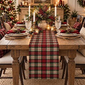 May include: A rectangular table runner featuring a red, green, and cream plaid pattern centered on a wooden dining table. The table is set with white plates, clear textured glassware, gold cutlery, and a centerpiece of pine branches and red berries.