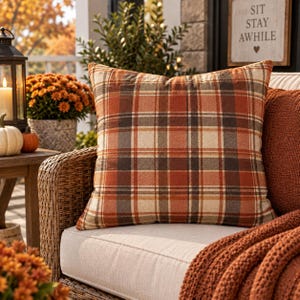 May include: A square throw pillow featuring a plaid pattern in shades of burnt orange, cream, brown, and dark gray. The fabric has a visible woven texture, and the pillow is positioned on a light-colored cushion next to a textured orange blanket.