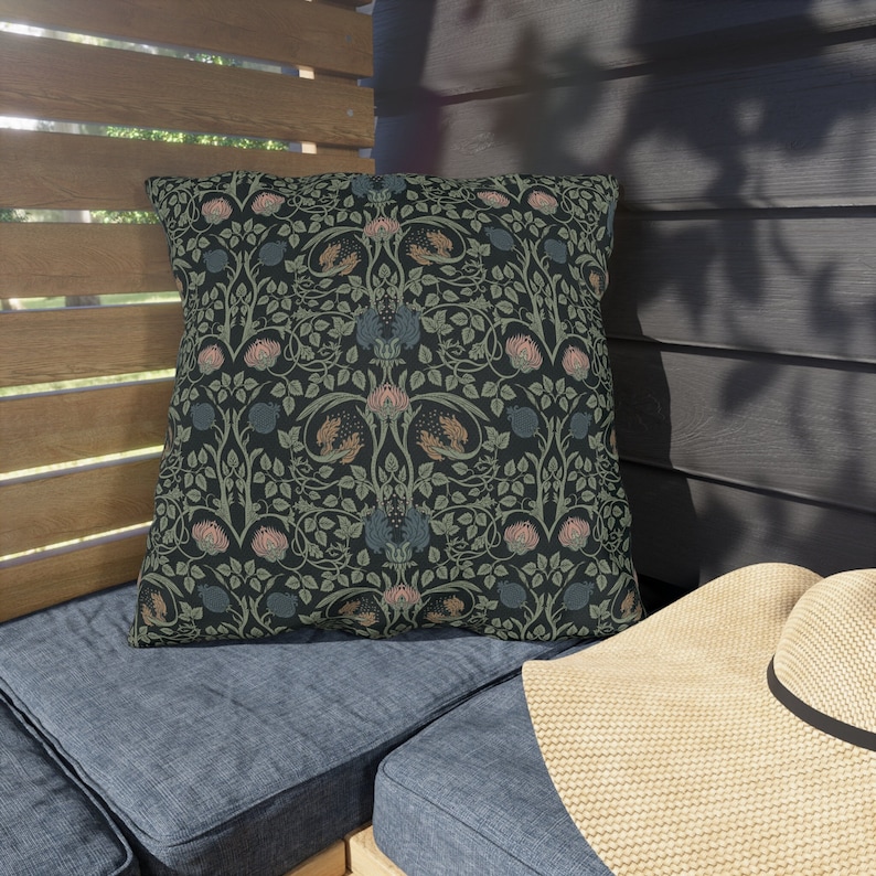 May include: Square decorative pillow with a dark background and a floral pattern in shades of green, pink, and blue. The pillow is placed on a blue cushion, with a straw hat in the foreground.