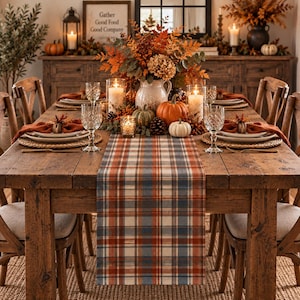 May include: A rustic wooden dining table set for a meal, featuring a plaid table runner in shades of orange, blue, and cream. The centerpiece includes a ceramic pitcher filled with autumn foliage, pumpkins, and lit candles in glass holders.
