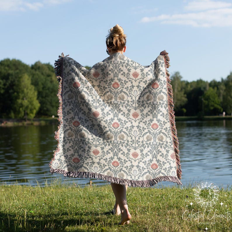 May include: A woman wearing a woven throw blanket with a floral pattern in shades of pink, green, and white. The blanket has a fringed edge and is draped over her shoulders. She is standing in front of a lake with trees in the background.