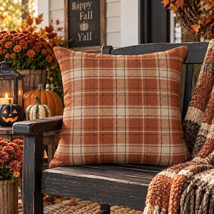 May include: A square throw pillow featuring a plaid pattern in shades of burnt orange, cream, and dark brown. The fabric has a textured appearance, and the pillow is positioned on a dark wooden bench, showcasing a classic woven textile design.