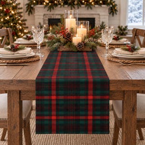 May include: A rectangular table runner featuring a classic plaid pattern in deep forest green, black, and vibrant red. The fabric lies flat across a wooden dining table, centered beneath a holiday centerpiece with three white pillar candles.