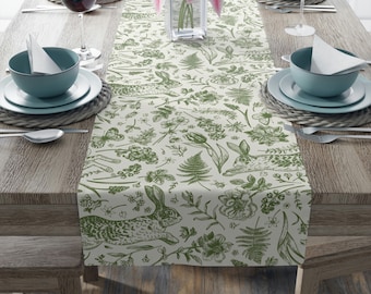 May include: A green and white table runner with a repeating pattern of rabbits, flowers, and ferns. The runner is on a wooden table with place settings on either side.
