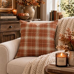 May include: A square throw pillow featuring a burnt orange, cream, and dark brown plaid pattern. The fabric has a visible woven texture, and the pillow is positioned on a light-colored upholstered chair, adding a warm, seasonal aesthetic to the space.