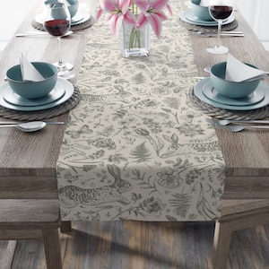 May include: A beige table runner with a green floral and rabbit pattern. The runner is laid out on a wooden table with two place settings on each side.