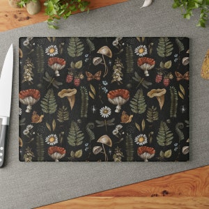 May include: A black cutting board with a repeating pattern of mushrooms, ferns, dragonflies, and flowers. The design is in shades of green, brown, red, and white.
