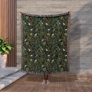 May include: A woven throw blanket with a dark background and a repeating pattern of green, white, and brown leaves and flowers. The blanket has a fringe border.