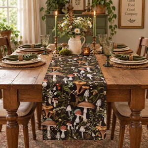 May include: A dark-colored table runner featuring a detailed botanical print of various mushrooms, ferns, and wildflowers. The runner is centered on a wooden dining table set with white plates, green napkins, and green glassware.