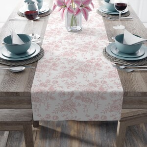 May include: A dining table set for a meal, featuring a white and pink floral patterned table runner. The table is set with blue bowls, white plates, silverware, and wine glasses. A vase of pink lilies sits in the center.