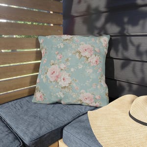 May include: A square throw pillow with a floral pattern of pink roses and green leaves on a light blue background. The pillow is placed on a blue cushion, with a straw hat in the foreground.
