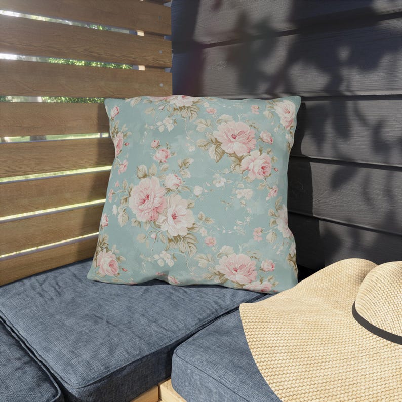 May include: A square throw pillow with a floral pattern of pink roses and green leaves on a light blue background. The pillow is placed on a blue cushion, with a straw hat in the foreground.