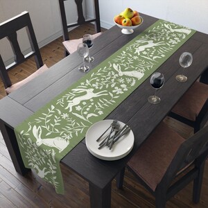May include: A green table runner with a white floral and bunny design. The runner is on a dark wood table with two chairs and a bowl of fruit.