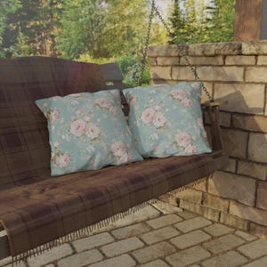 May include: Two square throw pillows with a floral pattern of pink roses on a light blue background rest on a wooden swing. The swing has a brown plaid blanket. The scene is outdoors with a brick wall and trees in the background.