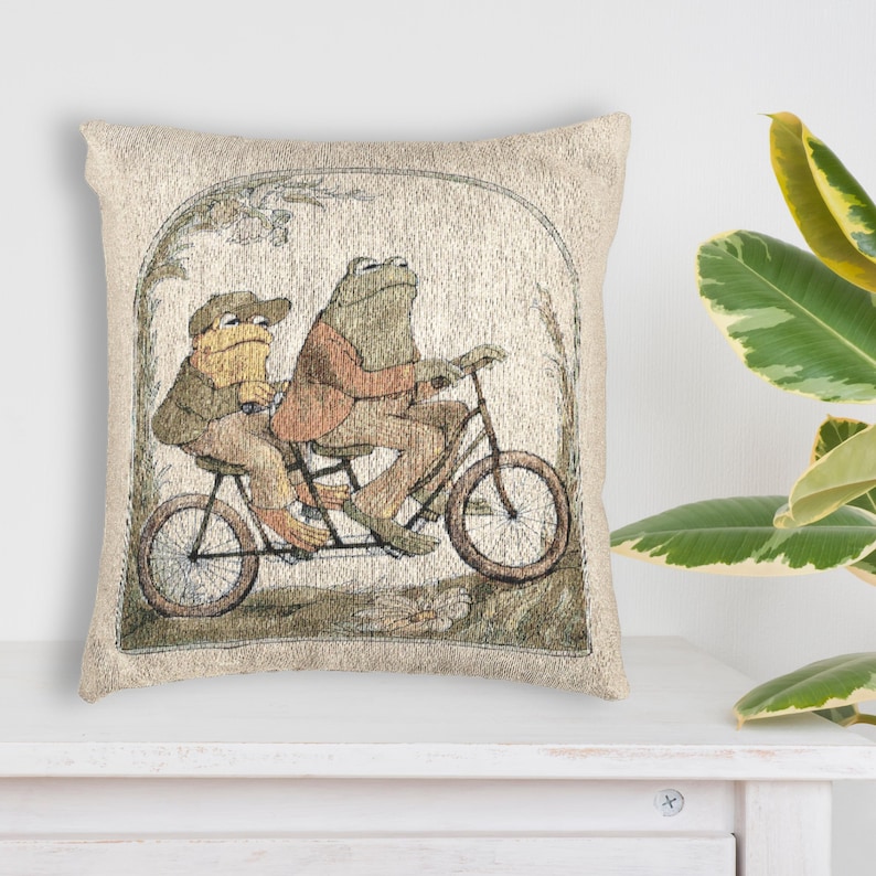 May include: A square throw pillow featuring a woven tapestry design of two frogs riding a tandem bicycle. The frogs are depicted in shades of brown and green, wearing jackets and hats, set within an arched frame against a light beige background.