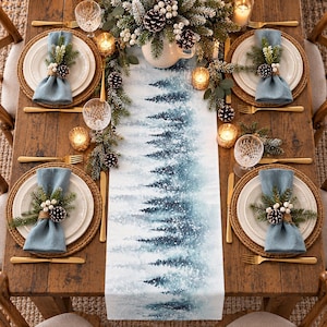 May include: A dining table setting featuring a white and blue table runner with a snowy pine forest print. Four place settings include woven chargers, white plates, blue napkins with pine and berry napkin rings, gold cutlery, and glassware.
