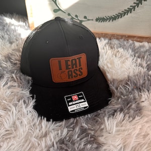 May include: A black trucker hat with a brown leather patch that says "I EAT ASS" with a peach emoji. The hat is made by Richardson 112 and is adjustable.