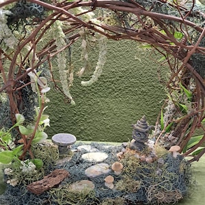 May include: A miniature fairy garden scene with a stone table and chairs, a small lantern, and mushrooms nestled in moss and greenery. The garden is surrounded by a twisted vine archway.
