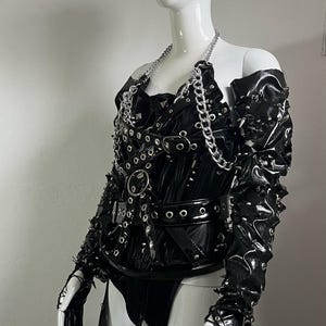 May include: A black leather corset with silver hardware, including chains, buckles, and eyelets. The corset has a pointed collar and a long skirt with a split in the front. The corset is worn on a white mannequin.