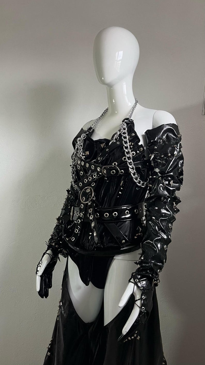 May include: A black leather corset with silver hardware, including chains, buckles, and eyelets. The corset has a pointed collar and a long skirt with a split in the front. The corset is worn on a white mannequin.