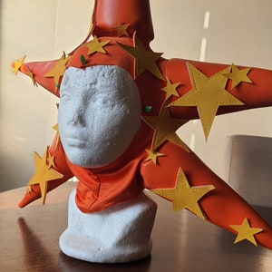 Może przedstawiać: Jasny pomarańczowy, gwiazdkowaty kapelusz z żółtymi filcowymi gwiazdami. Kapelusz jest wykonany z filcu i ma spiczasty wierzchołek. Kapelusz jest noszony na białej głowie z pianki styropianowej.