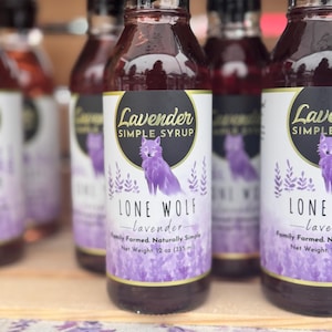 K&ouml;nnte beinhalten: Eine Reihe von Glasflaschen, gef&uuml;llt mit lila Fl&uuml;ssigkeit, mit den Etiketten "Lavender Simple Syrup" und "Lone Wolf - lavender - Family Farmed. Naturally Simple. Net Weight: 12 oz (355 ml)". Die Flaschen zeigen eine Wolfs-Illustration und einen lila-wei&szlig;en Aquarell-Hintergrund.