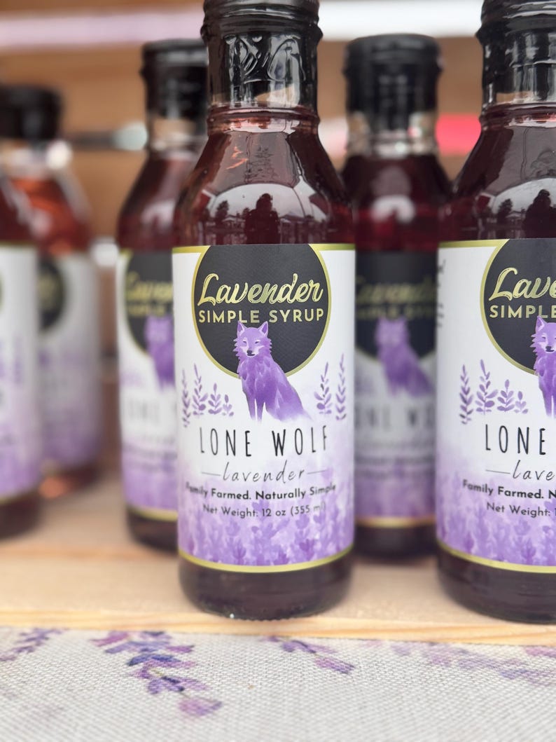 K&ouml;nnte beinhalten: Eine Reihe von Glasflaschen, gef&uuml;llt mit lila Fl&uuml;ssigkeit, mit den Etiketten "Lavender Simple Syrup" und "Lone Wolf - lavender - Family Farmed. Naturally Simple. Net Weight: 12 oz (355 ml)". Die Flaschen zeigen eine Wolfs-Illustration und einen lila-wei&szlig;en Aquarell-Hintergrund.