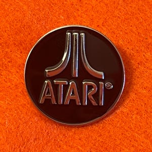 May include: A round, dark brown Atari pin with a silver border and the Atari logo in silver. The logo features three vertical lines above the word "ATARI". The pin is set against an orange background.