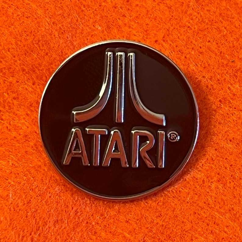 May include: A round, dark brown Atari pin with a silver border and the Atari logo in silver. The logo features three vertical lines above the word "ATARI". The pin is set against an orange background.