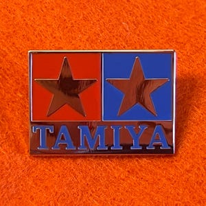 May include: A rectangular Tamiya pin with a red and blue color scheme. The pin features two stars and the word "TAMIYA" in blue lettering. The pin has a shiny, metallic finish.