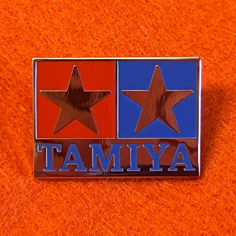 May include: A rectangular Tamiya pin with a red and blue color scheme. The pin features two stars and the word "TAMIYA" in blue lettering. The pin has a shiny, metallic finish.